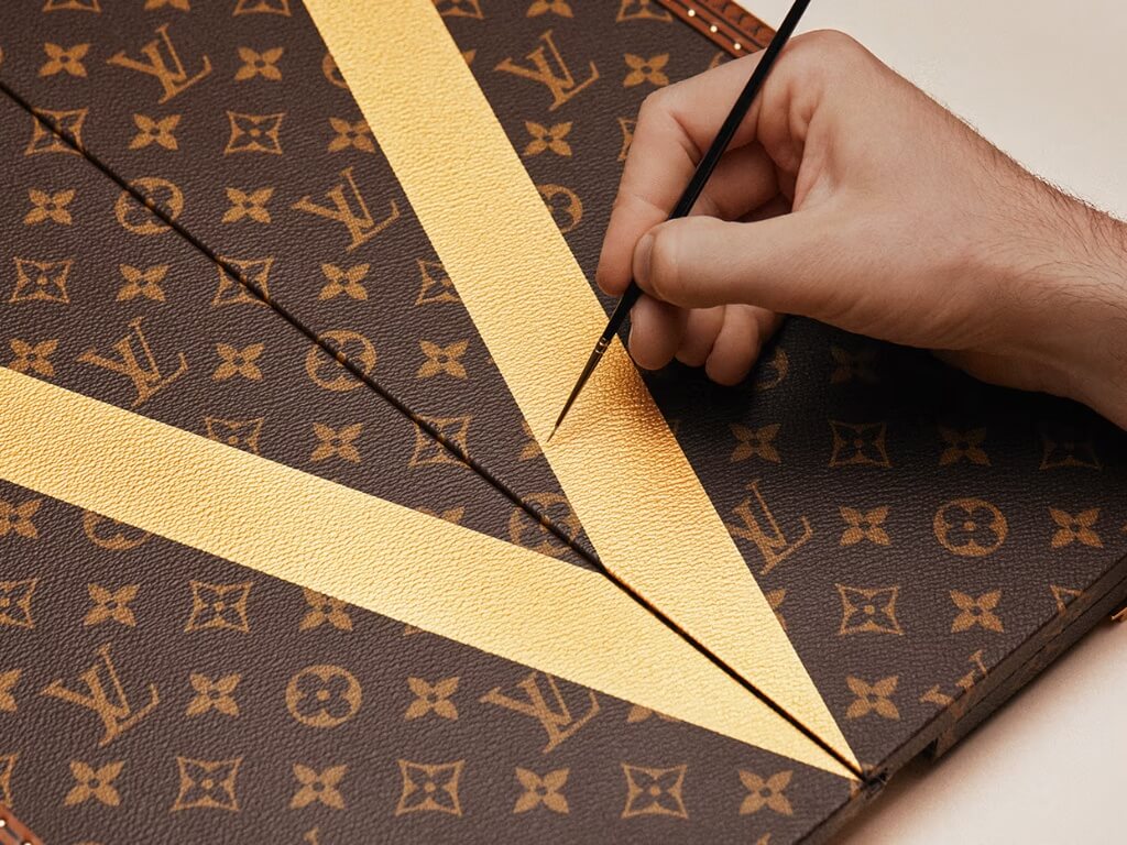 Λεπτομέρεια του Monogram καμβά και του χρυσού συμβόλου V στο Louis Vuitton FIFA World Cup 2026 Trophy Trunk.